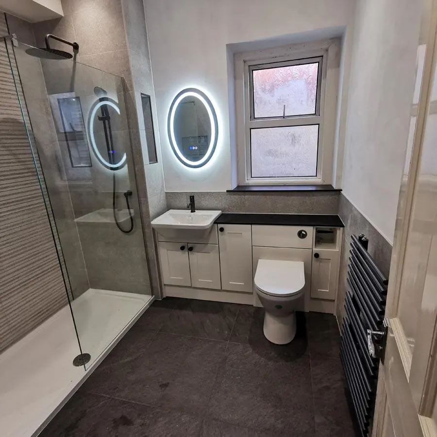 Bathroom with a walk-in shower, round lit mirror, white vanity unit and black towel radiator