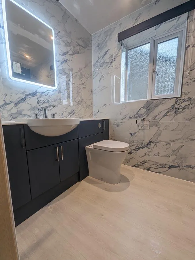 Navy vanity unit and toilet with marble-effect tiles and a lit mirror